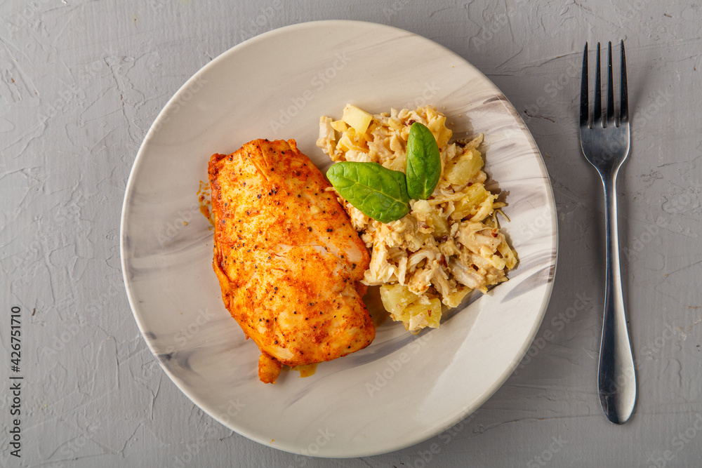 Baked chicken breast with salad on a plate next to a fork on a gray background. Baked chicken breast with salad on a plate next to a fork on a gray background.
