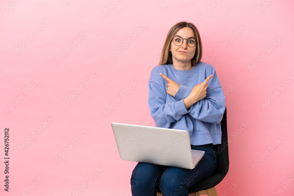 Naklejka premium Young woman sitting on a chair with laptop over isolated pink background pointing to the laterals having doubts