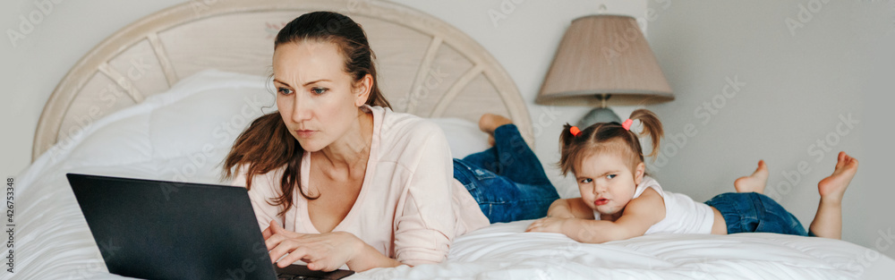 Work from home with kids children. Mother working on laptop in bedroom with child daughter toddler beside her. Funny candid authentic lifestyle family moment. Web banner header. Work from home with kids children. Mother working on laptop in bedroom with child daughter toddler beside her. Funny candid authentic lifestyle family moment. Web banner header.