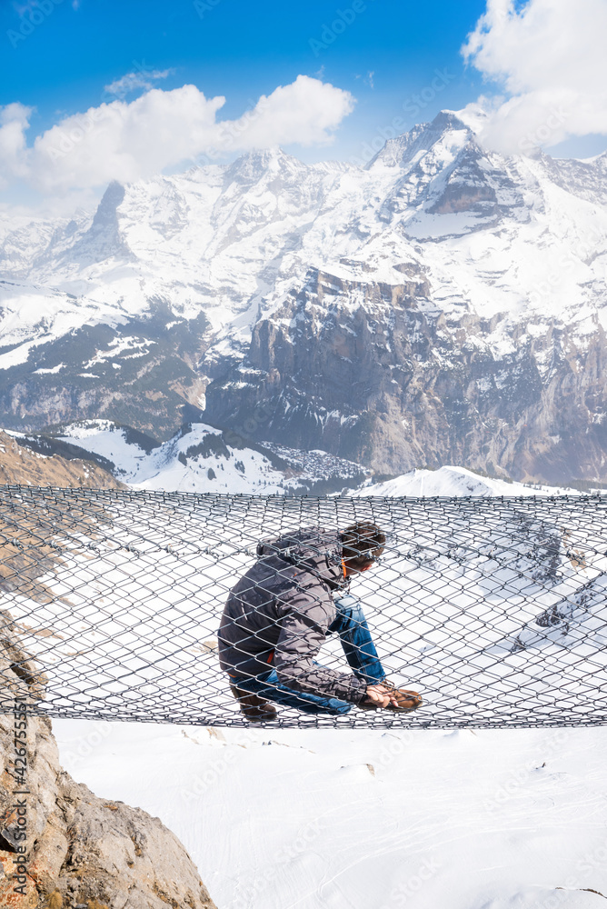 A young man climbs over a wire mesh at an altitude, in the background with the Swiss Alps.