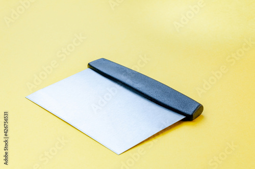Metal dough scraper knife over bright yellow surface background. Stainless steel flat dough scraper with plastic handle