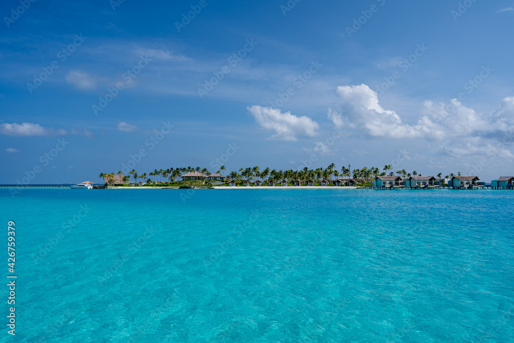 Blick auf eine Insel der Malediven, mit Palmen, Strand, Overwater Bungalows und dem kristallklaren und türkisblauen Wasser