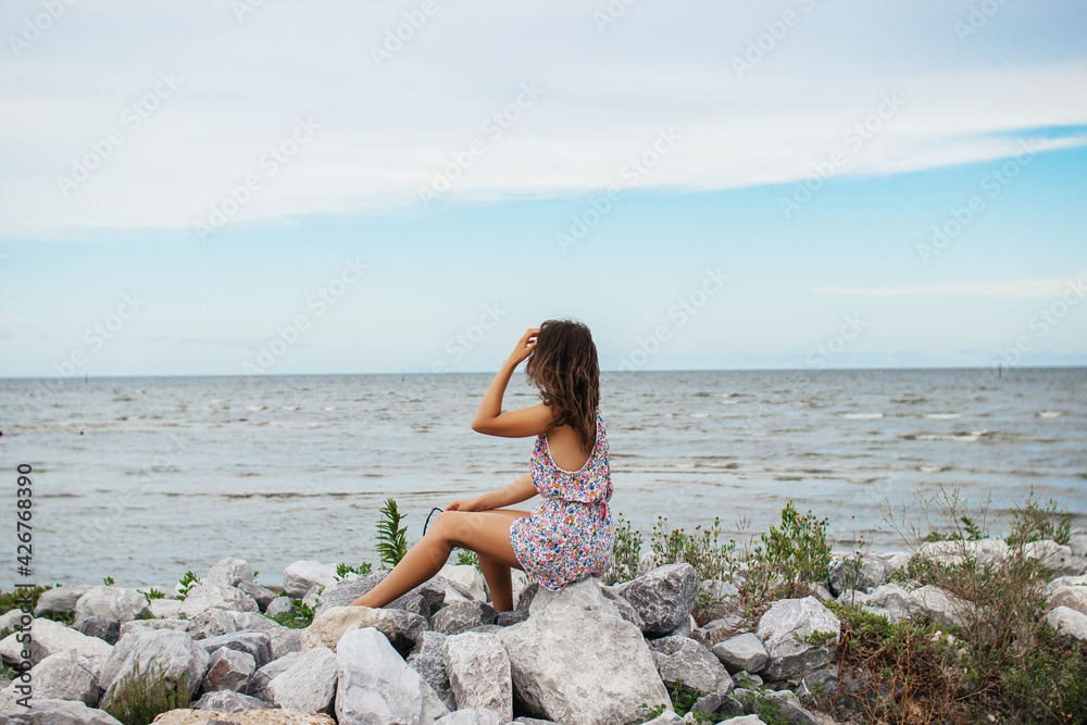 A beautiful young woman sits on a stone shore and looks at the ocean. Summer background. A beautiful young woman sits on a stone shore and looks at the ocean. Summer background.