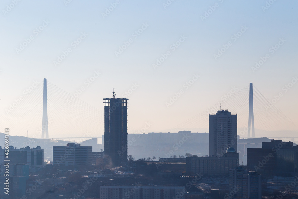 The silhouette of the Russian bridge across the Eastern Bosphorus is in dense fog. Russian bridge in Vladivostok against the background of residential buildings under construction.