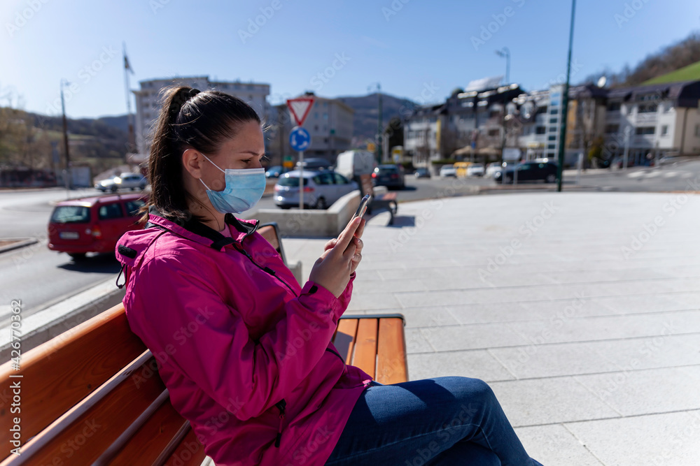 Fototapeta premium A young woman sits in a park bench alone and uses a smartphone. Virus pandemic times