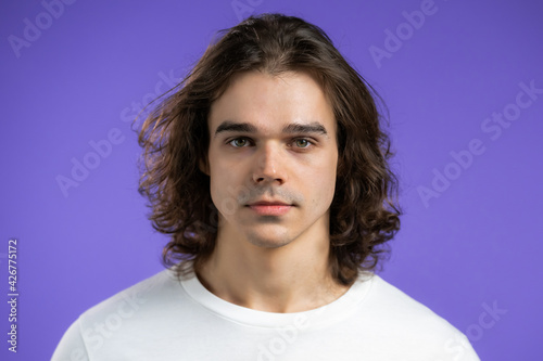 Portrait of serious european man looking to camera. Young handsome guy with long hairdo on violet studio background.