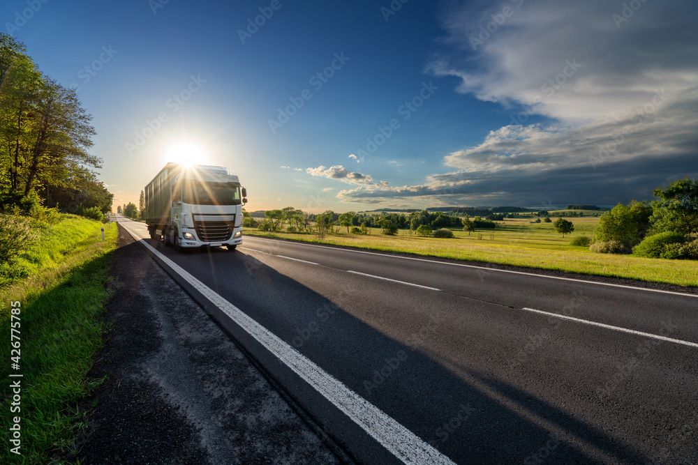 White truck driving on the asphalt road in rural landscape in the rays of the sunset