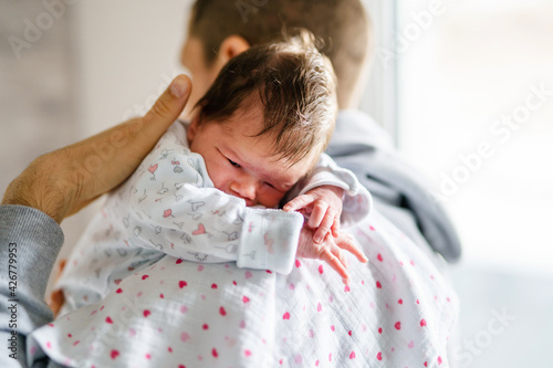 Back view of father holding his newborn baby over the shoulder while standing by the window in day at home - sleepy infant in position for burping new life and parenthood concept