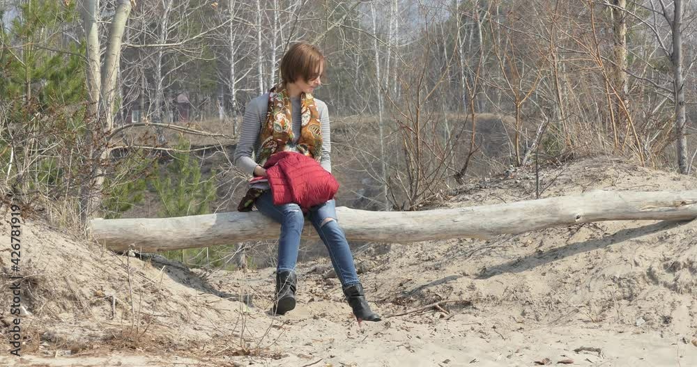 A woman in self-isolation in jeans sits on a log on a sandy beach against the background of spring trees that are not yet covered with leaves and looks around. A woman in self-isolation in jeans sits on a log on a sandy beach against the background of spring trees that are not yet covered with leaves and looks around.