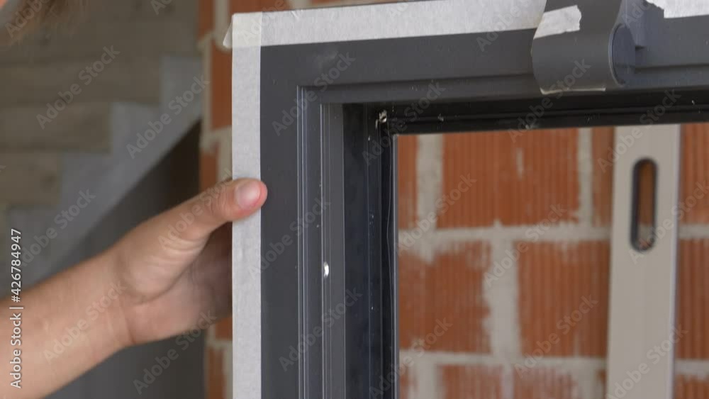 CLOSE UP: Handyman uses white adhesive tape to protect the black window frame before mounting it at a house under construction. Unrecognizable worker applies protective tape around a window frame.