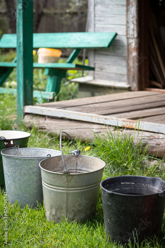 Many different buckets stand in a row to collect rainwater. Buckets of water stand on the grass in the garden next to the house. Village concept. Old wooden house and bench in the background.