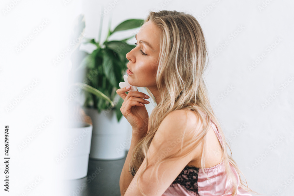 Blonde smiling woman 35 year clean fresh face and hands with long hair does facial massage with stone scraper in underwear at bathroom Blonde smiling woman 35 year clean fresh face and hands with long hair does facial massage with stone scraper in underwear at bathroom