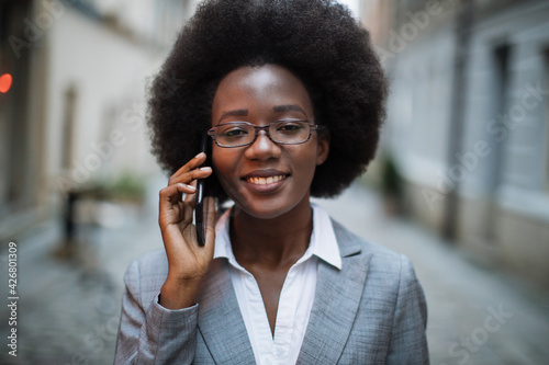 Portrait of smiling african lady in stylish suit and eyeglasses using modern smartphone for conversation outdoors. Concept of business, people and technology.