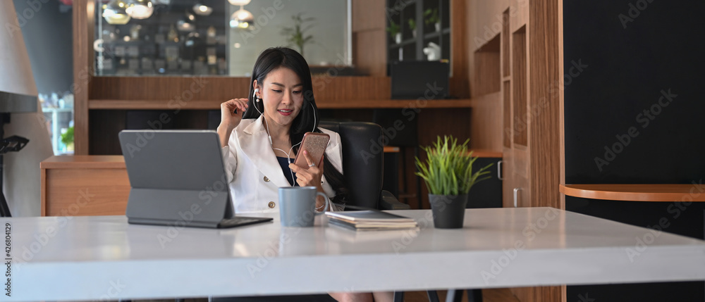 Casual businesswoman in earphone and looking at smart phone while sitting at her workplace. Casual businesswoman in earphone and looking at smart phone while sitting at her workplace.