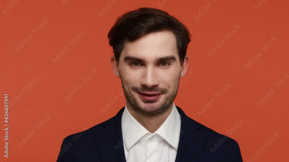 Handsome young brunette bearded man dresses suit winking and looking at camera with optimistic smile, flirting, expressing optimism. Indoor studio shot isolated on orange background. Handsome young brunette bearded man dresses suit winking and looking at camera with optimistic smile, flirting, expressing optimism. Indoor studio shot isolated on orange background.