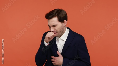 Seasonal flu. Unhealthy bearded man in official suit coughing covering mouth with hand, choke allergy or influenza symptoms, feeling unwell. Indoor studio shot isolated on orange background
