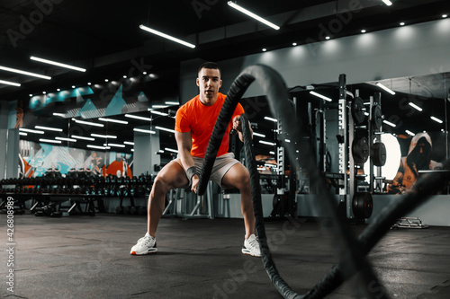 An energetic sportsman is doing heavy cardio cross-fit training with battle ropes in an indoor gym with a black background and a big mirror. Functional training, sports lifestyle