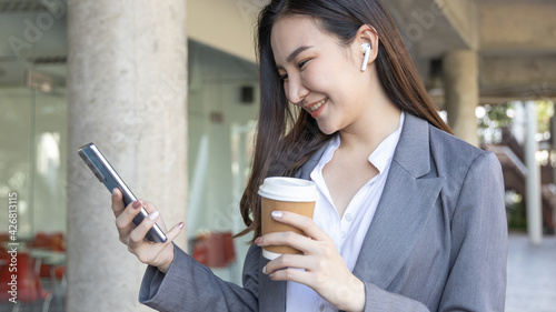 Young Asian woman wearing a gray suit is holding a phone and a paper coffee mug, Business women use smartphones to communicate by chatting or talking with business partners, Online media, VDO call.