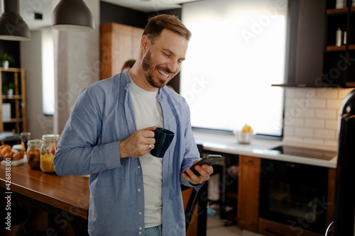 Cheerful young man is drinking a coffee in kitchen and watching something fun on his phone.