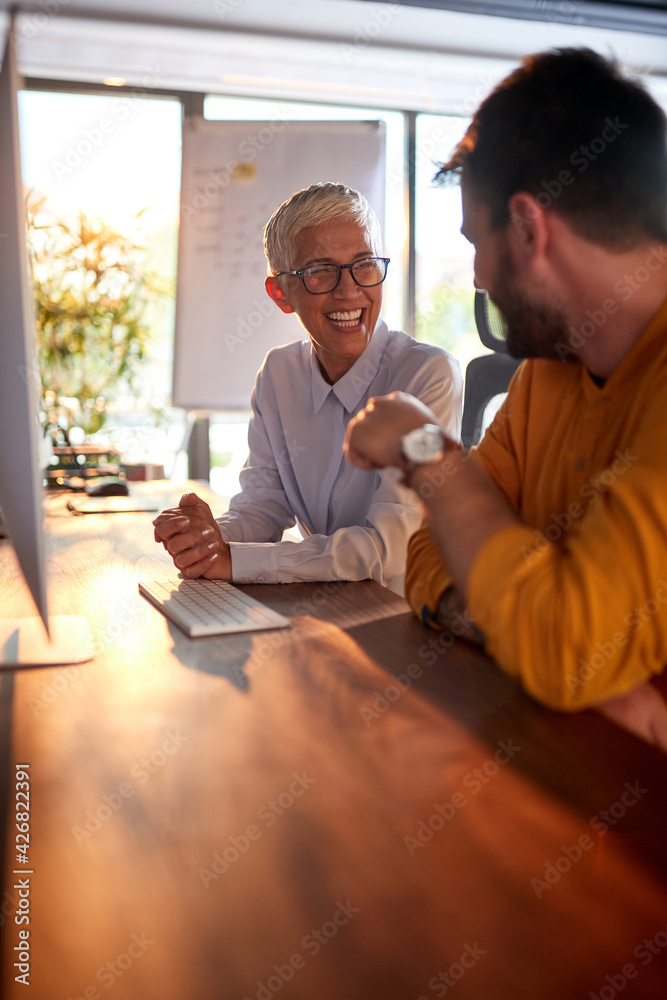 An elderly business woman has a good time with her young male colleague while they working at the desk in the office. Business, office, job - obrazy, fototapety, plakaty 