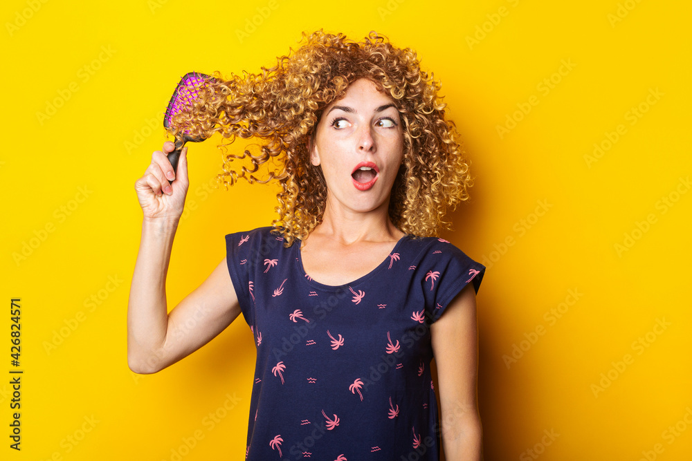 surprised young woman combing tangled curly hair on yellow background surprised young woman combing tangled curly hair on yellow background