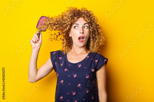 surprised young woman combing tangled curly hair on yellow background