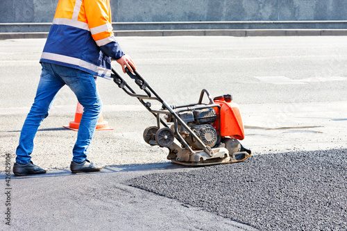 A road worker compacts the asphalt with a gasoline vibratory plate compactor.