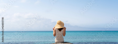 Rear view woman wearing a straw hat sitting on a rock looking at the translucent sea. Horizontal banner or header © Pintau Studio