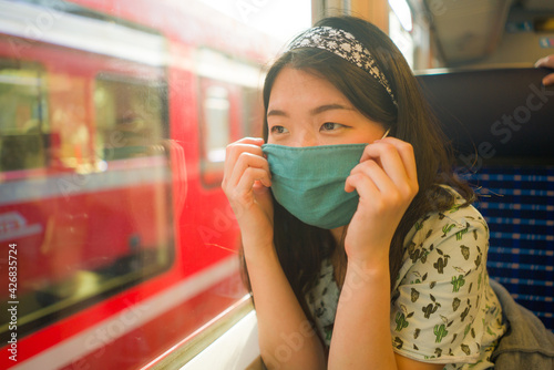 train travel in times of covid19 - young happy and cute Asian Korean woman in face mask traveling on railcar looking landscape through the window enjoying holiday getaway