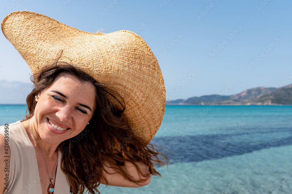 Happy smiling woman portrait with closed eyes wearing a straw hat in the beach with wind that ruffles hair. beautiful translucent sea on background.