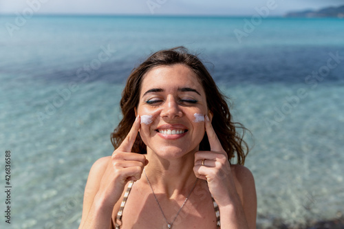 Close-up of young woman applying sunscreen in her face in a sunny day at the beach. Translucent blue clear water on background