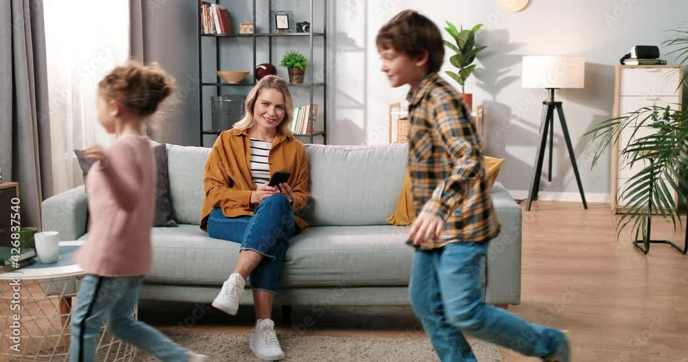 Cheerful adorable Caucasian small children boy and girl running and playing in apartment while their mom sitting on sofa at home typing on smartphone browsing online and smiling, family concept