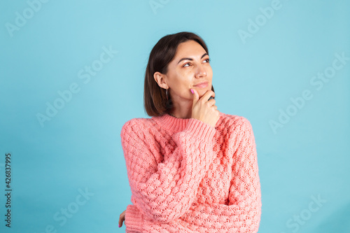Young brunette in warm pink sweater isolated on blue background looks to the side thoughtfully with a sweet smile