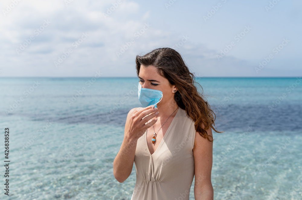 Portrait of young woman standing against blue clear water on the sea. Female taking off a surgical protective face mask Portrait of young woman standing against blue clear water on the sea. Female taking off a surgical protective face mask