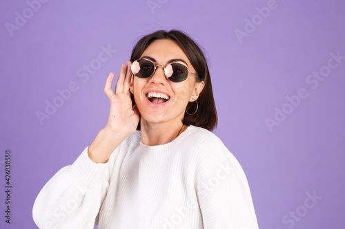 Young brunette in white casual sweater isolated on purple background wearing sunglasses happy smile positive moving