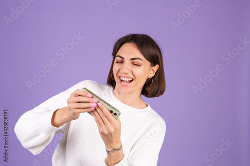 Young brunette in white casual sweater isolated on purple background happy overjoyed playing games on mobile phone