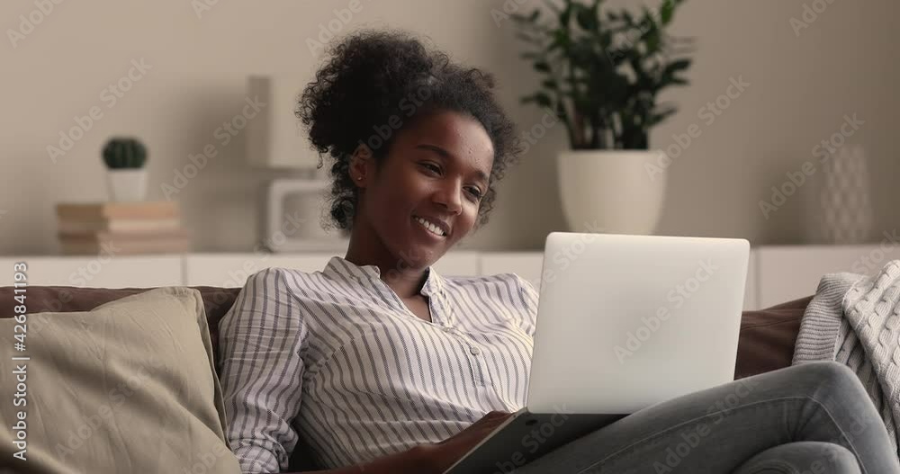 Relaxed millennial smiling african mixed race woman looking at laptop screen, watching funny movie film or pleasant video photo content in social network, shopping in internet store or web surfing. Relaxed millennial smiling african mixed race woman looking at laptop screen, watching funny movie film or pleasant video photo content in social network, shopping in internet store or web surfing.