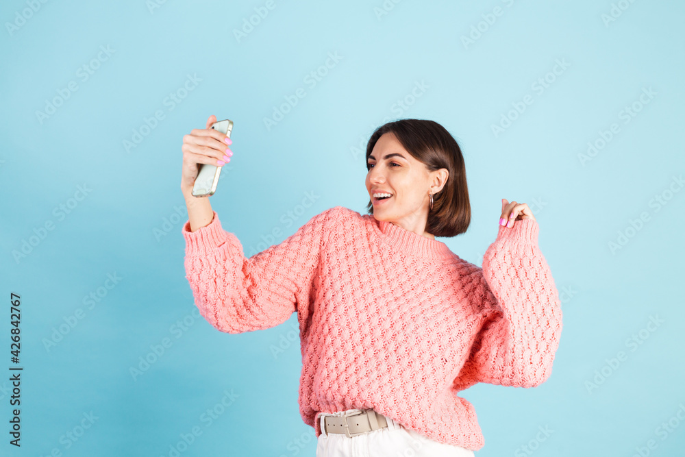 Young brunette in pink sweater isolated on blue background smiling and looking at camera while taking selfie photo Young brunette in pink sweater isolated on blue background smiling and looking at camera while taking selfie photo