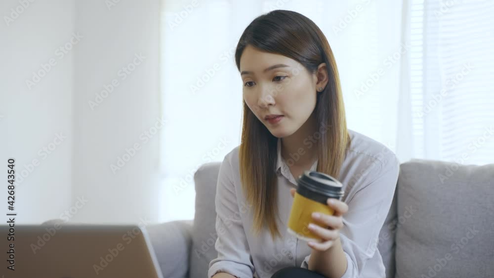 Young Asian business woman sitting on sofa using laptop reading online newspaper and drinking coffee in living room in morning. Young Asian business woman sitting on sofa using laptop reading online newspaper and drinking coffee in living room in morning.