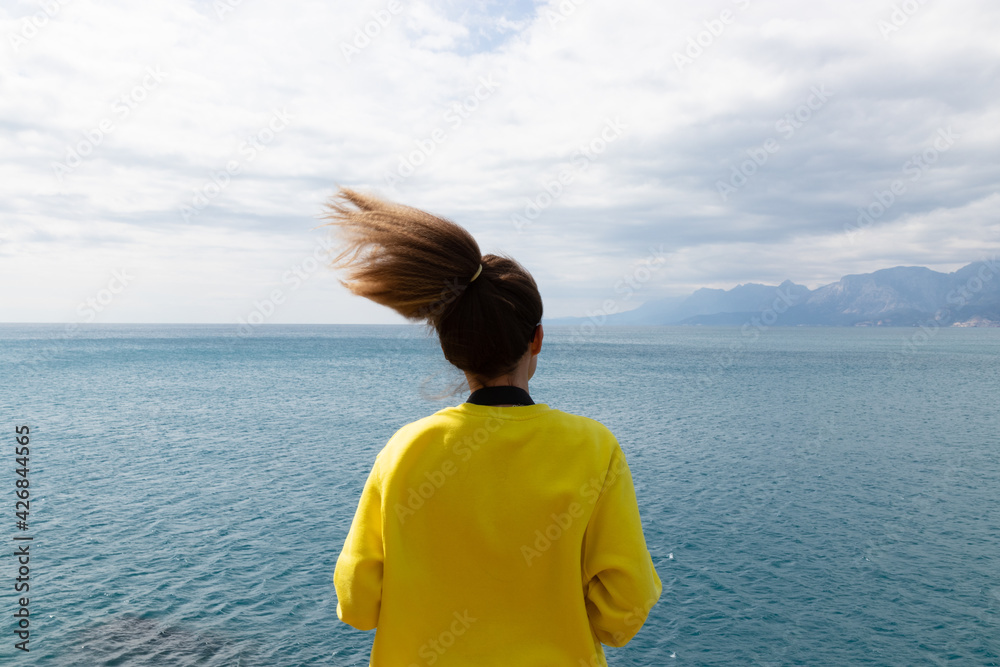 Back photography of young woman wearing yellow long sleeve stands in front of the sea under blue sky and white clouds.