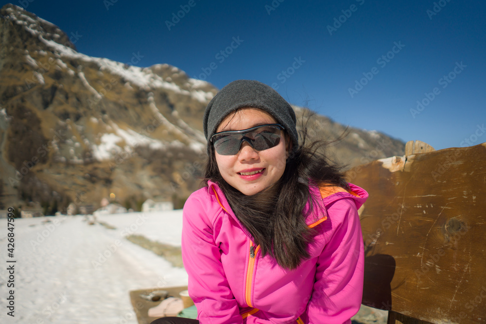 freezing Winter holidays - young happy and beautiful Asian Korean woman on bench at frozen lake landscape surrounded by snow mountains enjoying Swiss Alps getaway