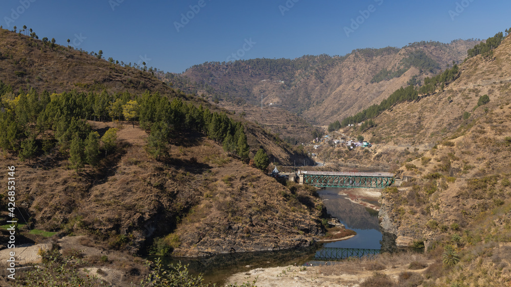 View of a river flowing through a narrow valley with rocky mountains on the sides and a bridge on top of it in the Kumaun region of UttaraKhand India on 12 January 2021

