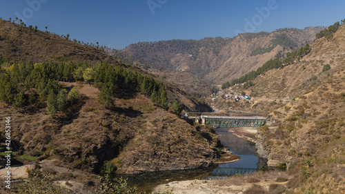 View of a river flowing through a narrow valley with rocky mountains on the sides and a bridge on top of it in the Kumaun region of UttaraKhand India on 12 January 2021 