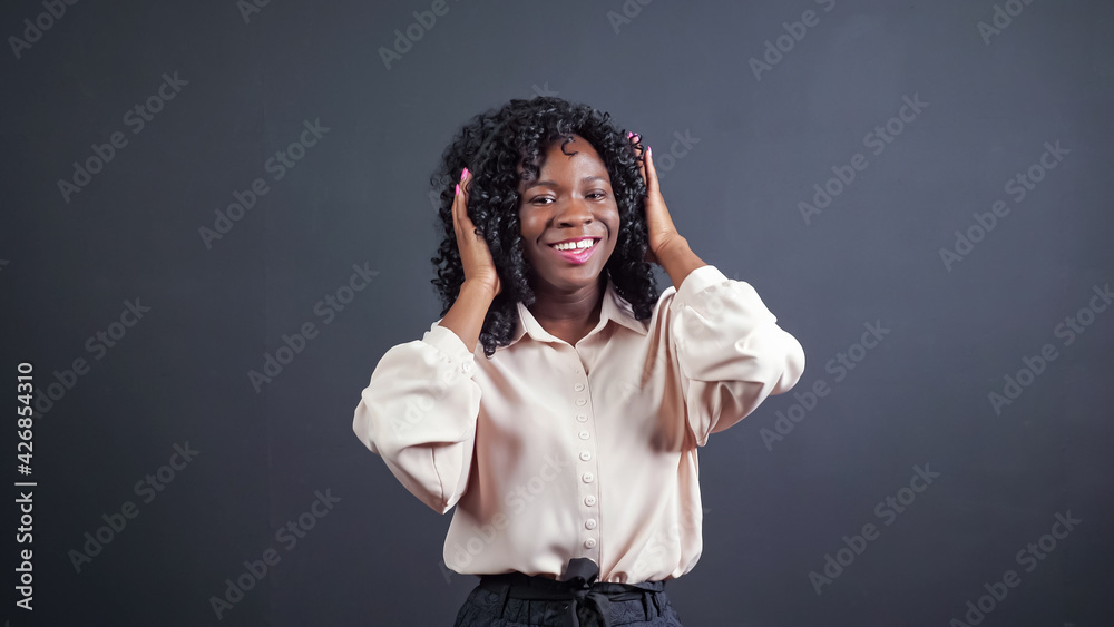 Cheerful young African-American lady with modern headphones in loose blouse dances posing for camera on black background in studio