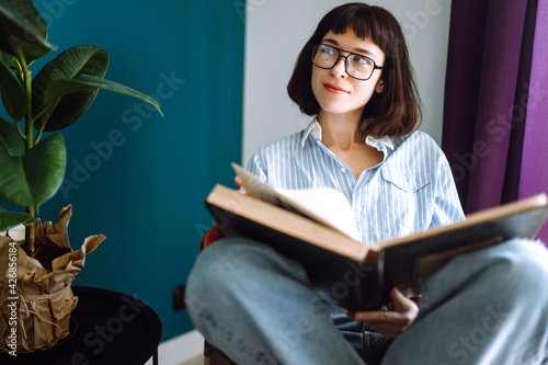 A young woman reads a book at home, sitting  in her living room. Concept of rest, relaxation, comfort, learning, reading, business.