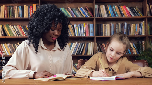 Professional individual Afro-American tutor with long curly hair dictates text to blonde schoolgirl and junior student writes sitting in library