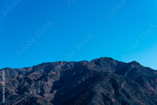 Green and brown forested mountain top under a blue sky.