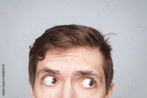Man's eyes looking up on the light background. Empty place for a text or object. Close-up shot of shocked young man with round eyes, top half head. Eyes looking on right side