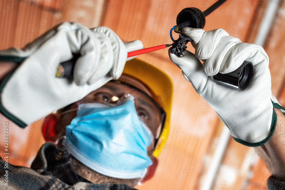 Electrician worker at work replaces the lamp holder protected by helmet, safety goggles and gloves; wear the surgical mask to prevent the spread of Coronavirus. Covid-19 Pandemic Prevention. Electrician worker at work replaces the lamp holder protected by helmet, safety goggles and gloves; wear the surgical mask to prevent the spread of Coronavirus. Covid-19 Pandemic Prevention.