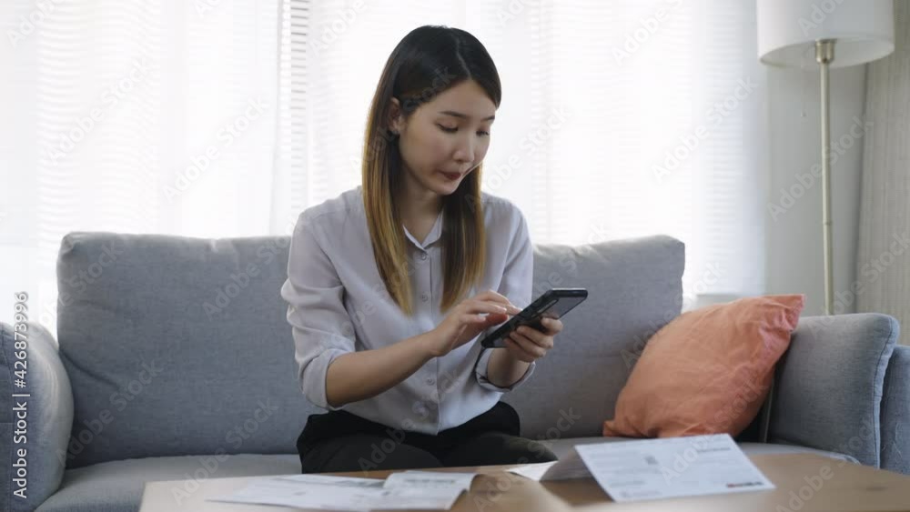 Stressed Asian woman sitting on the sofa looking at the paper bills calculating credit card invoice or unpaid taxes and bank account at home. Stressed Asian woman sitting on the sofa looking at the paper bills calculating credit card invoice or unpaid taxes and bank account at home.